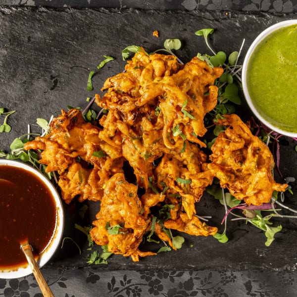 Crispy Indian pakora with green and brown dipping sauces on a dark slate board.