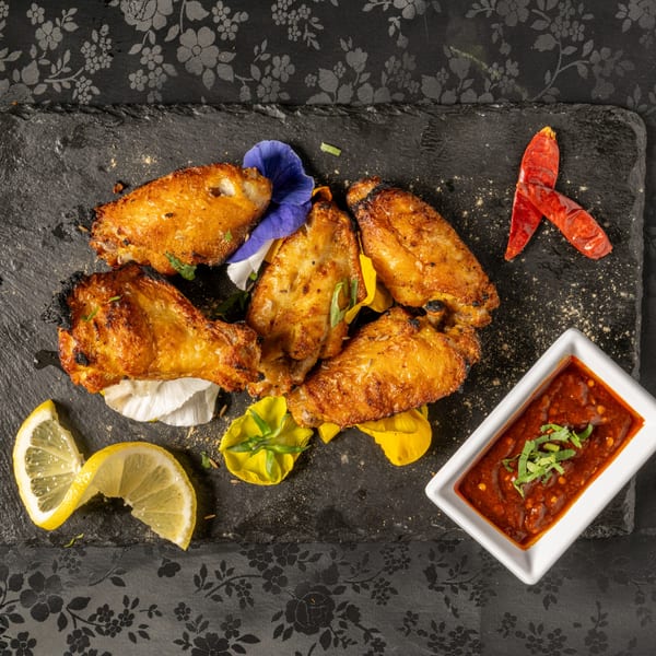 Golden-brown Indian chicken wings with lemon, dried chilies, and dipping sauce on a dark slate.