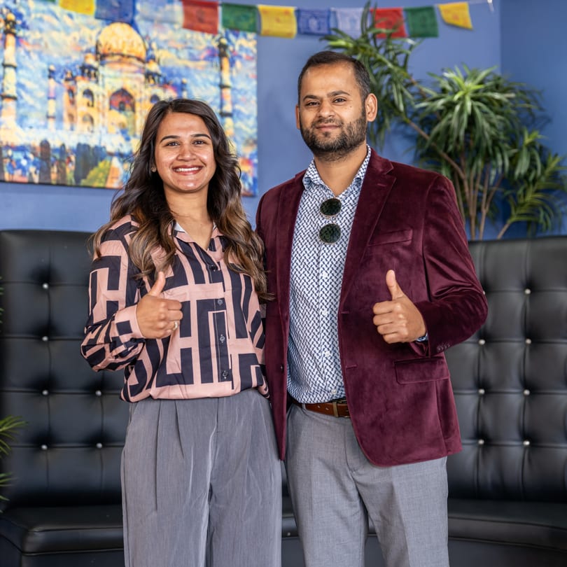 Two smiling people, a woman and a man, giving thumbs up in front of a Taj Mahal painting.