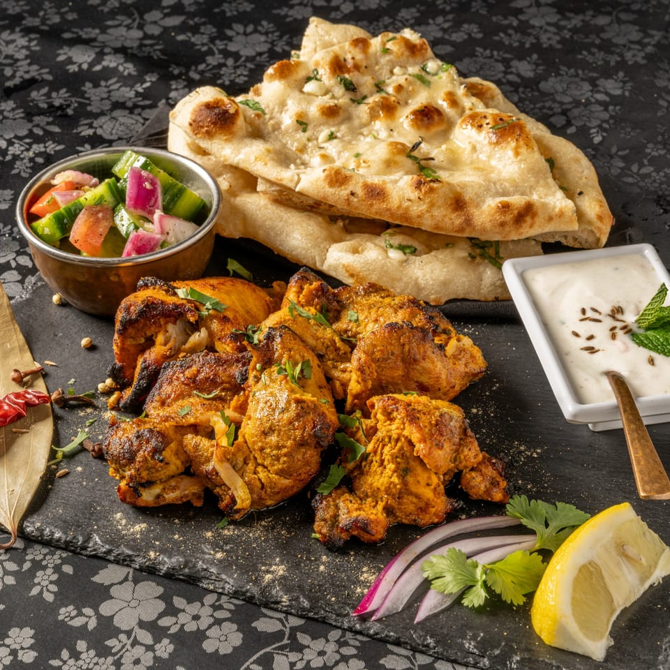 MOMO Curry's chicken tikka, naan bread, cucumber salad, and raita on a dark patterned tablecloth.