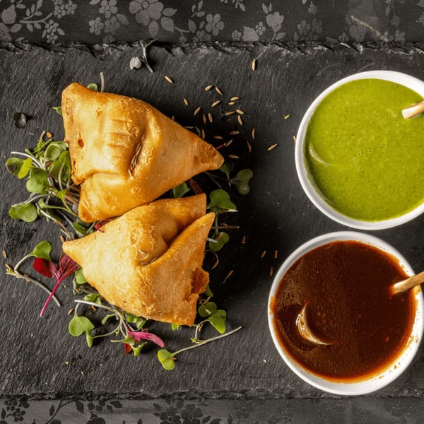 Two golden-brown samosas garnished with microgreens and cumin seeds, served with green and brown chutneys.
