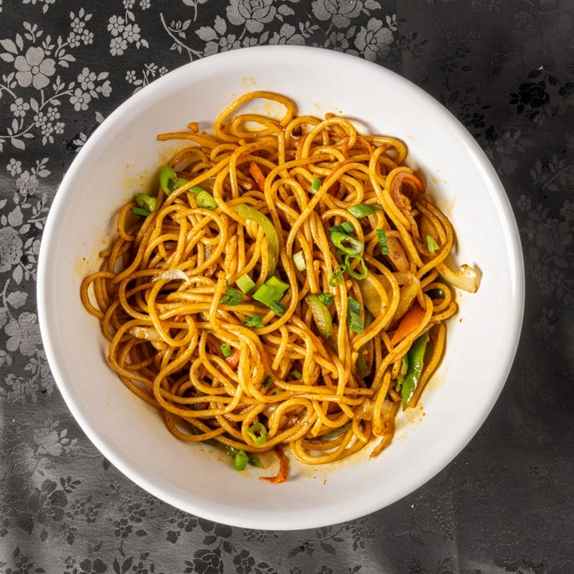 Overhead view of a white bowl filled with Indian noodles, vegetables, and green onions.
