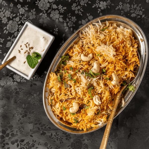 Indian biryani with cashews and mint in a silver dish, with raita on a black tablecloth.
