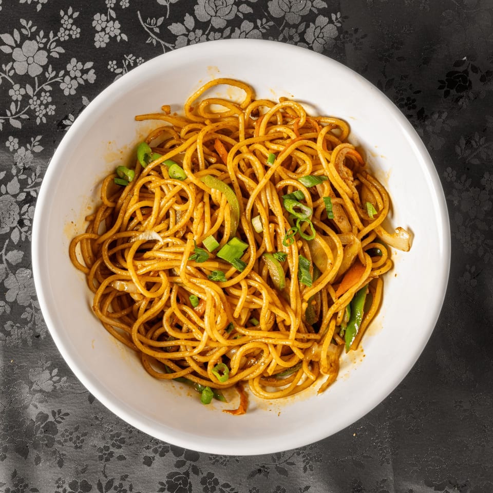 A white bowl filled with Indian-style noodles, green onions, and vegetables on a patterned tablecloth.