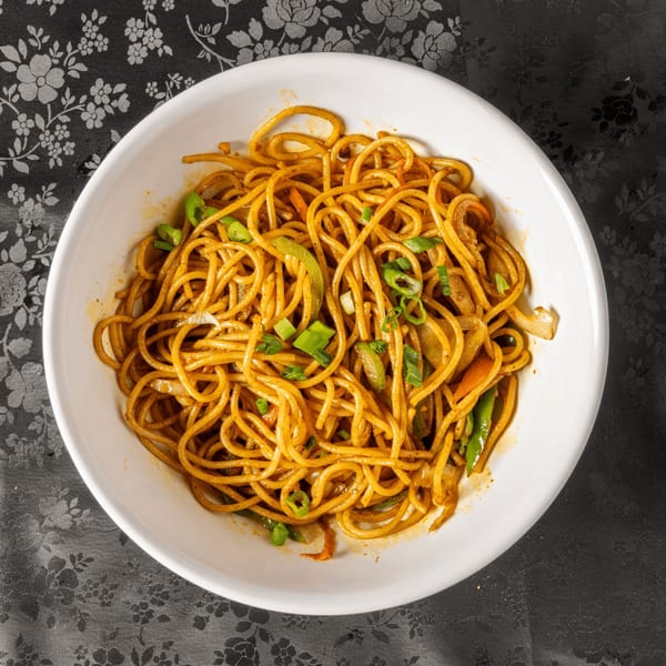 A white bowl filled with Indian noodles, green onions, and vegetables on a floral tablecloth.